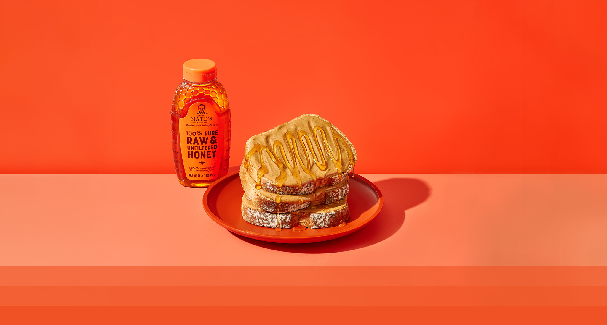 Bottle of Nate's Honey next to plate of toast with peanut butter and honey on orange background