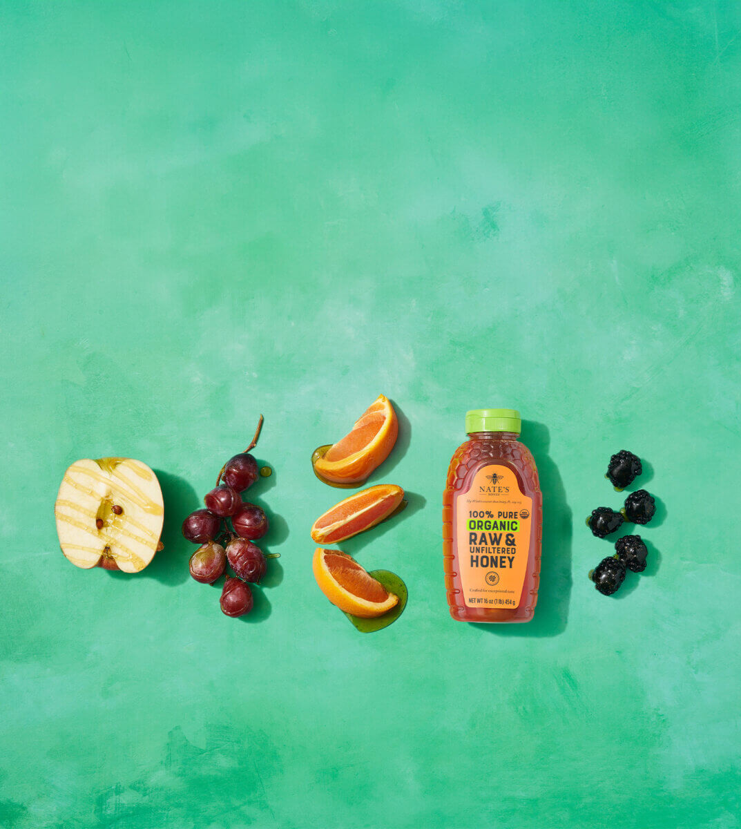 Nate's honey bottle with honey-drizzled fruits on green backdrop.