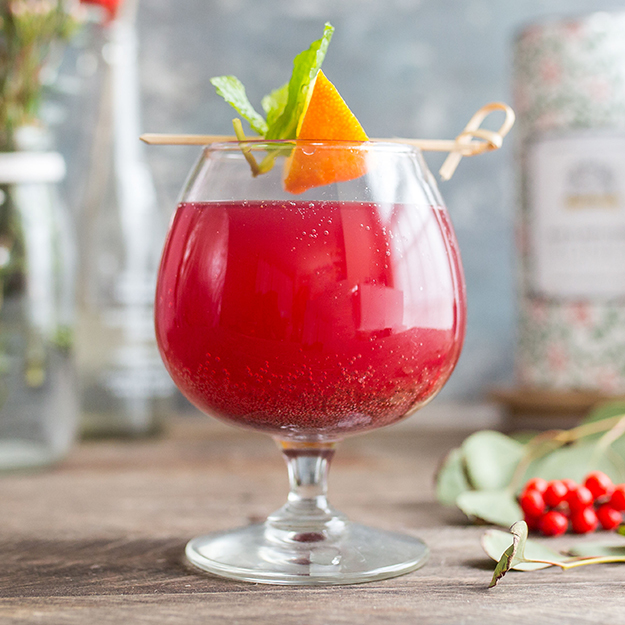 Blood orange drink in a stemmed glass garnished with an orange peel and mint, on a rustic table.