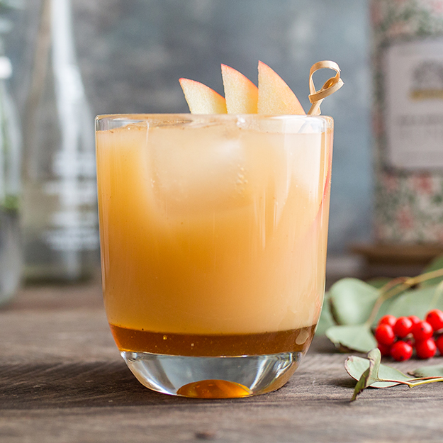 A golden cocktail with apple slice garnish in a clear glass, set against a rustic backdrop.