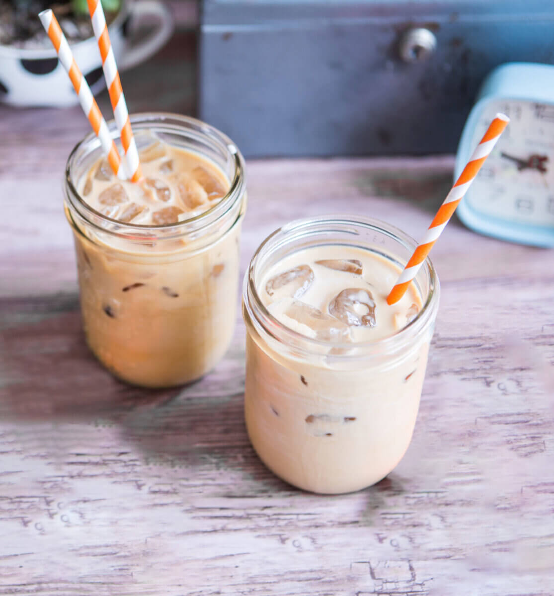 Two iced lattes with orange striped straws on a desk with laptop, clock, and keys.