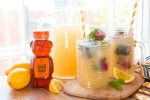 Lemonade in mason jar glasses and a pitcher, with a bottle of Nate's raw honey, on a wooden surface by a window.