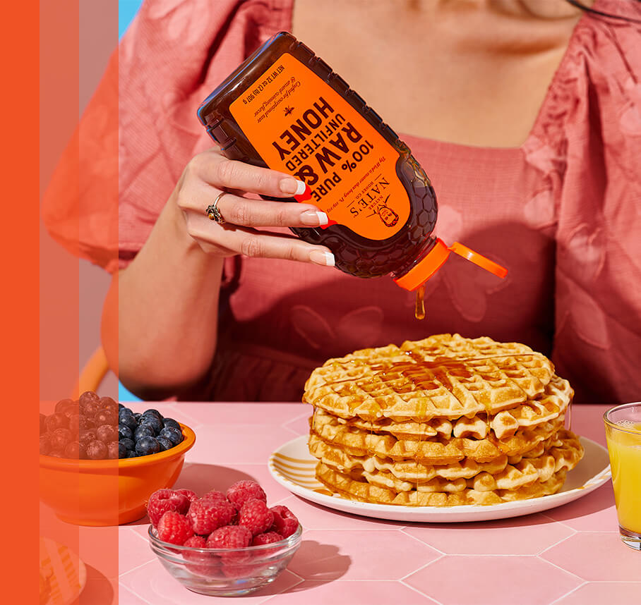 Woman enjoying stack of waffles by drizzling Nate's honey over top. Raspberries and blueberries in bowls next to waffles.