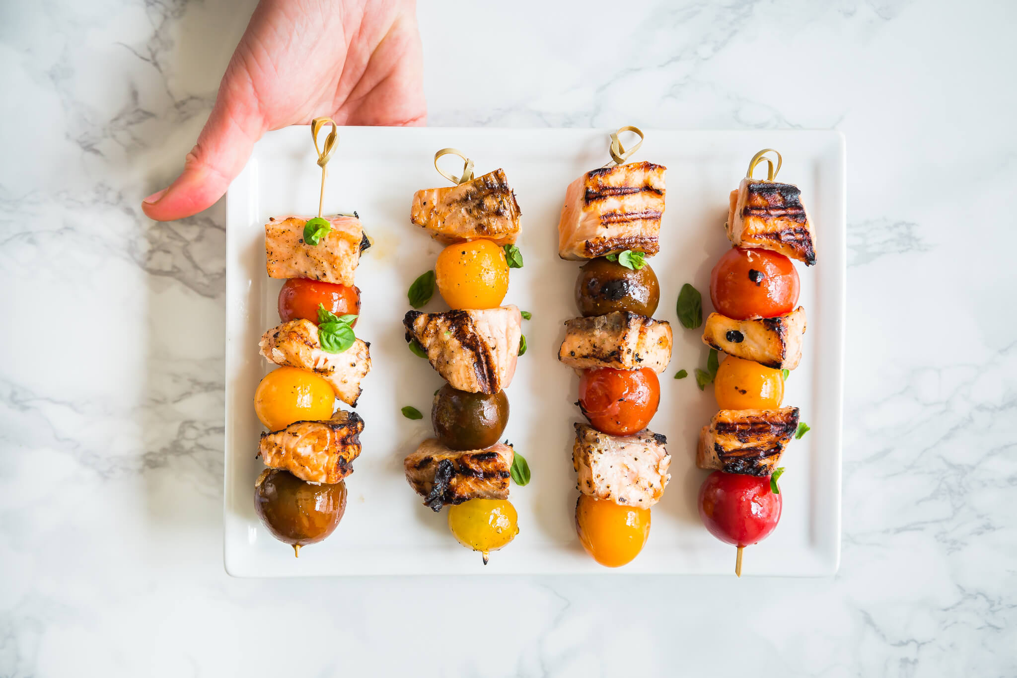 Marble table with hand holding white platter with Grilled Tomato Salmon Skewers