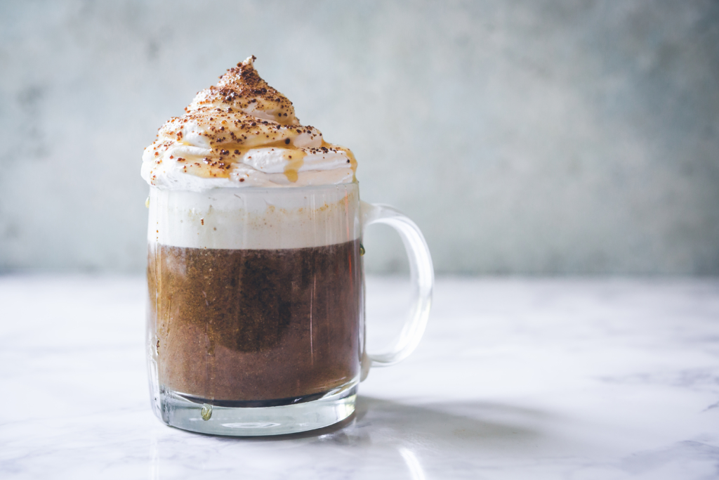 A glass mug of pumpkin latte topped with whipped cream, a honey drizzle and chocolate shavings sits on a marble surface.
