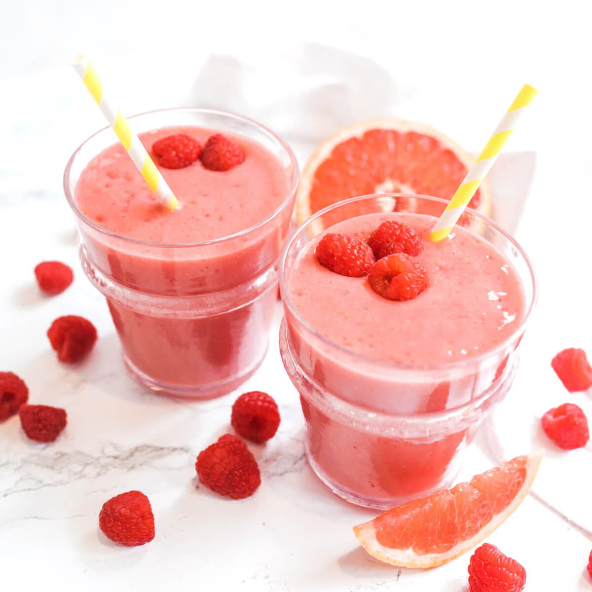 Glasses of raspberry smoothie with straws, fresh raspberries on top, and grapefruit slices on the side.