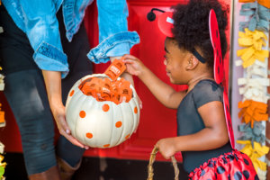A child in a ladybug costume reaching for a Nate's honey mini from a white pumpkin with orange spots, held by an adult.