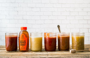 A row of assorted homemade sauces in jars with a bottle of Nate's honey, against a white brick wall. A row of assorted homemade sauces in jars with a bottle of Nate's honey, against a white brick wall.
