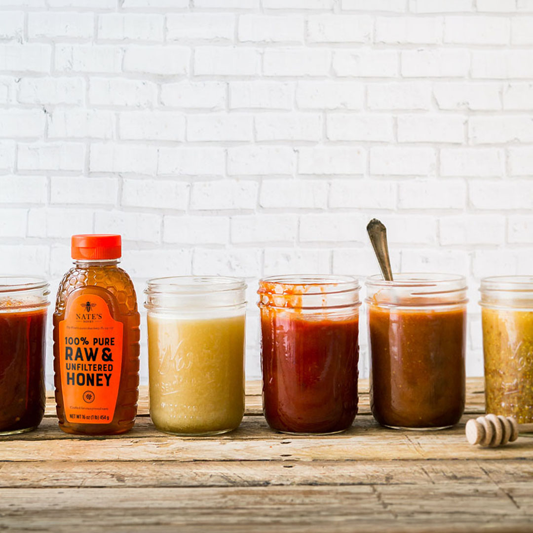 Assorted jars and a bottle of Nate's Honey on wooden surface with honey dipper and sauce.