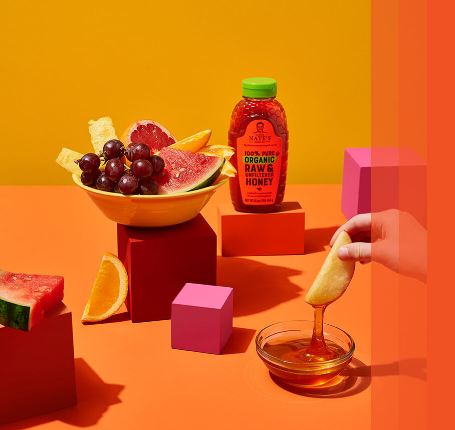 Fruit bowl next to Nate's honey bottle on an orange background. Apple in hand dipping into honey.
