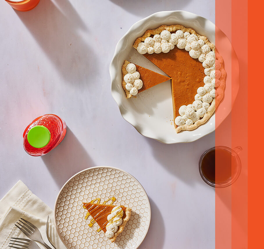 Pumpkin pie in dish topped with decorative whipped cream puffs lining the crust. One pie slice on plate drizzled with honey.