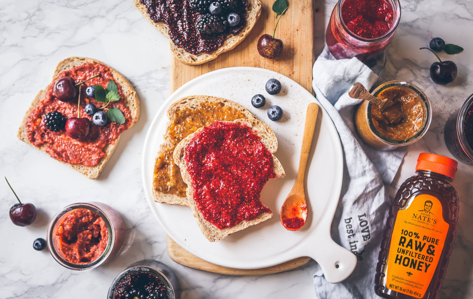 Breakfast spread with toast, fruit jam, berries and a bottle of Nate's Raw & Unfiltered Honey on a marble surface.