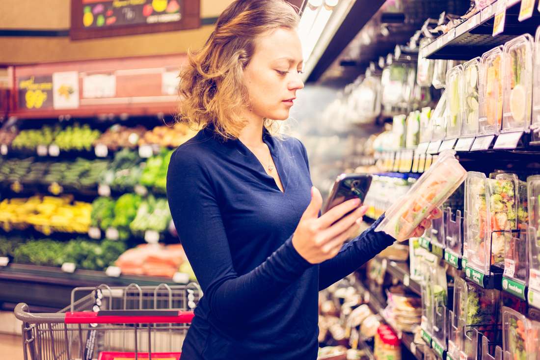 A person in a blue top is reading a label on a product in a supermarket aisle with a shopping cart nearby.