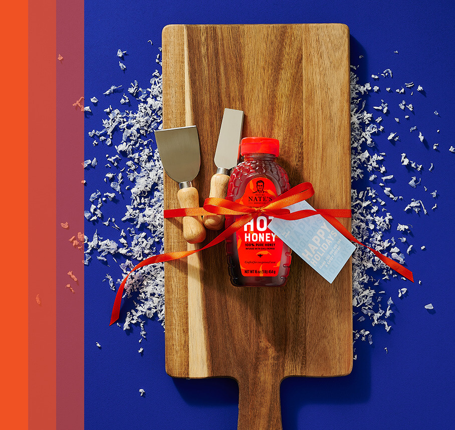 Wooden cutting board with Nate's hot honey bottle, serving tools wrapped with bow and gift tag decorated with snow confetti.