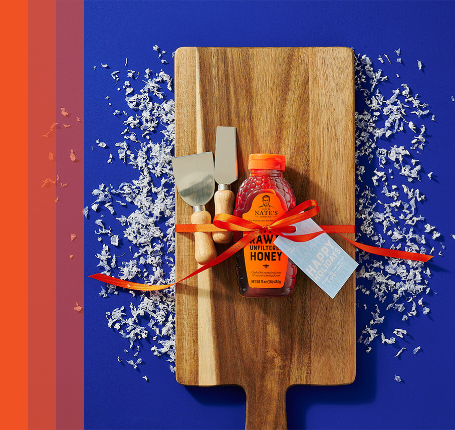 Wooden cutting board with Nate's honey bottle and serving tools wrapped with bow and gift tag decorated with snow confetti.