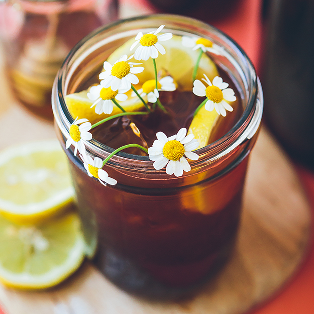 A refreshing glass of iced tea garnished with chamomile and lemon, with lemon slices and a warm-toned background.