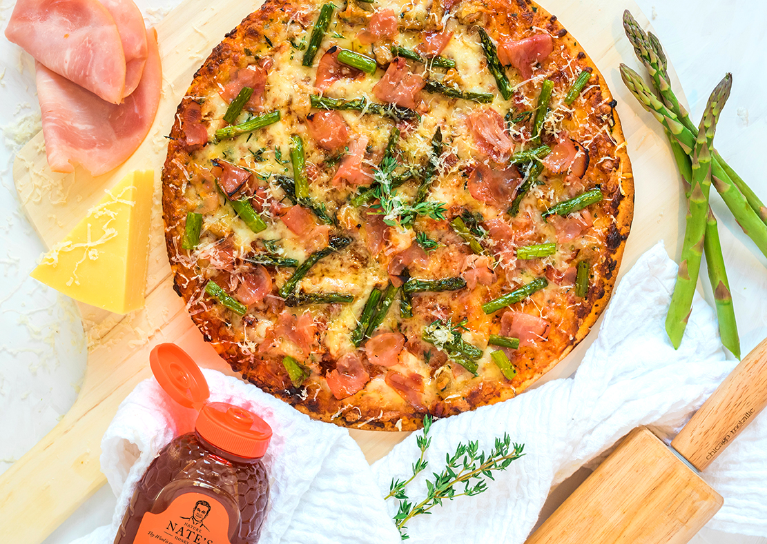 Pizza with asparagus and prosciutto, cheese and a bottle of Nate's honey on a white crinkled cloth.