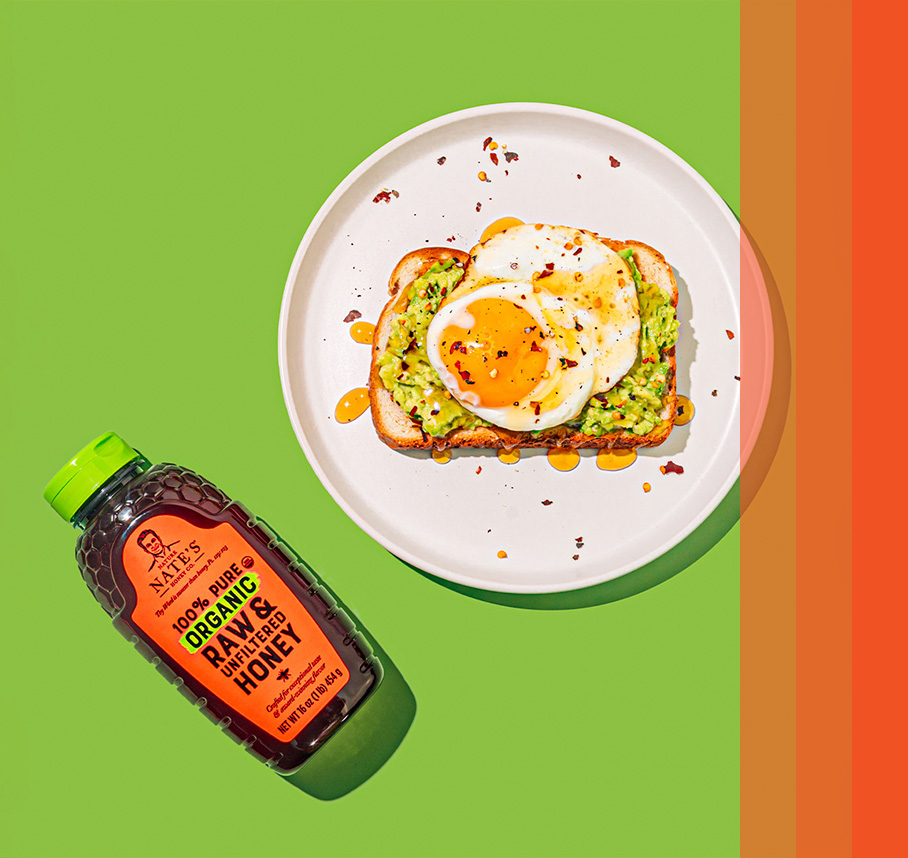Toast with avocado spread, sunny side up egg, red pepper flakes and honey drizzle next to Nate’s organic honey bottle.