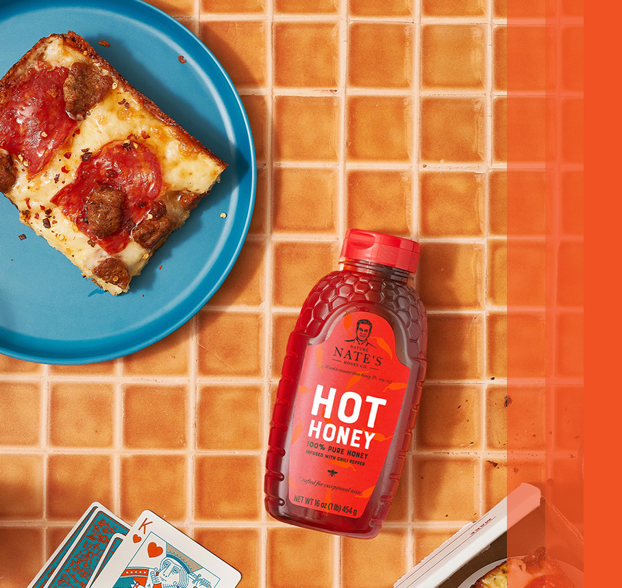 Nate's hot honey bottle next to a plate with pepperoni and sausage pizza slice and playing cards with a King face up.