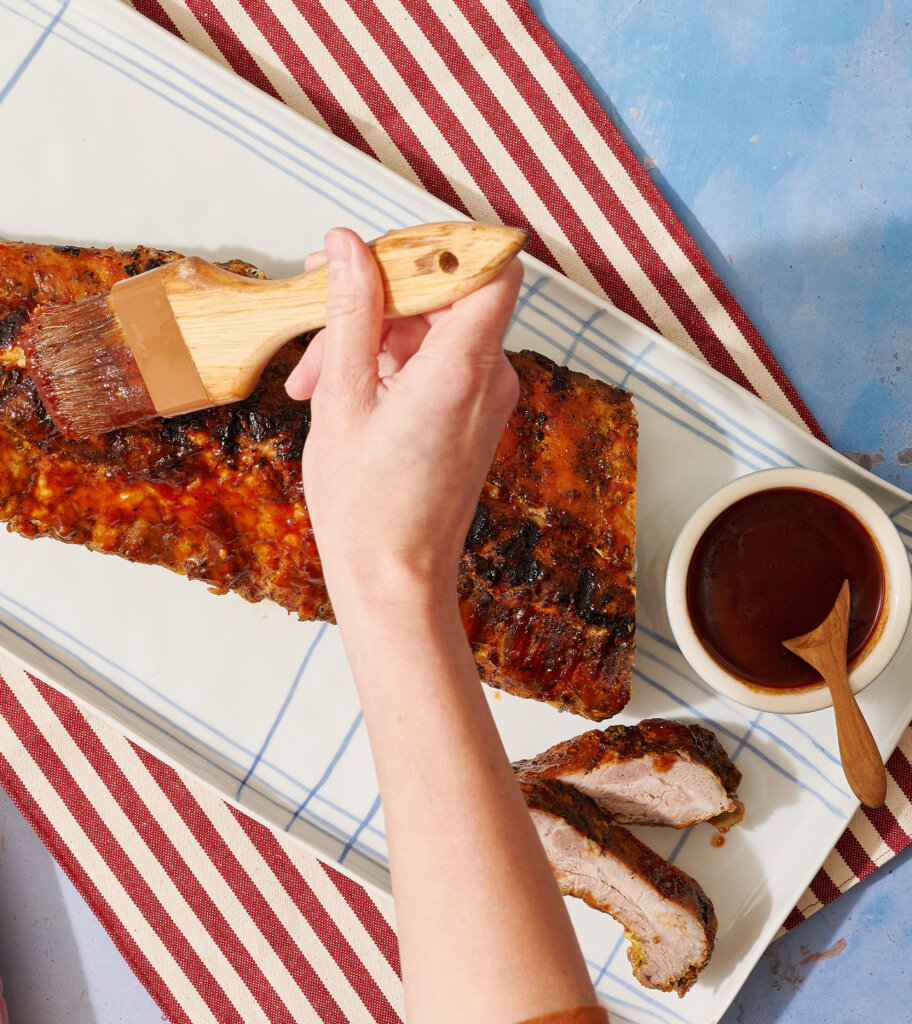 Glazed grilled pork ribs on plate with sauce, hand with basting brush, striped cloth.