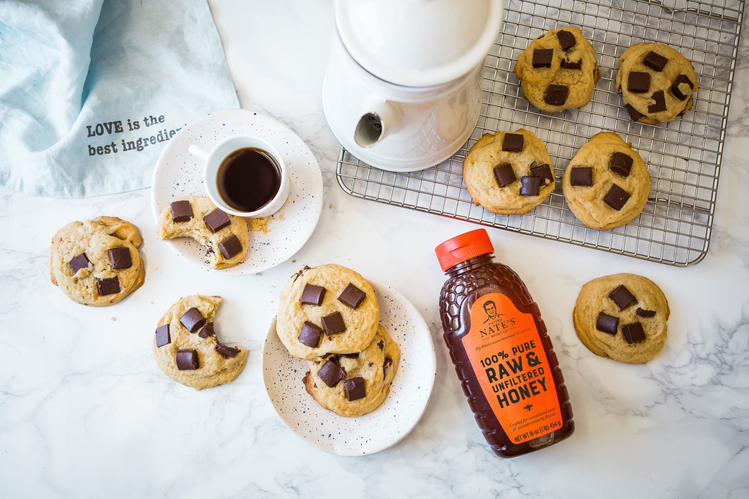 Chocolate chunk cookies with a bottle of Nate's honey, coffee and a napkin with the quote Love is the best ingredient.