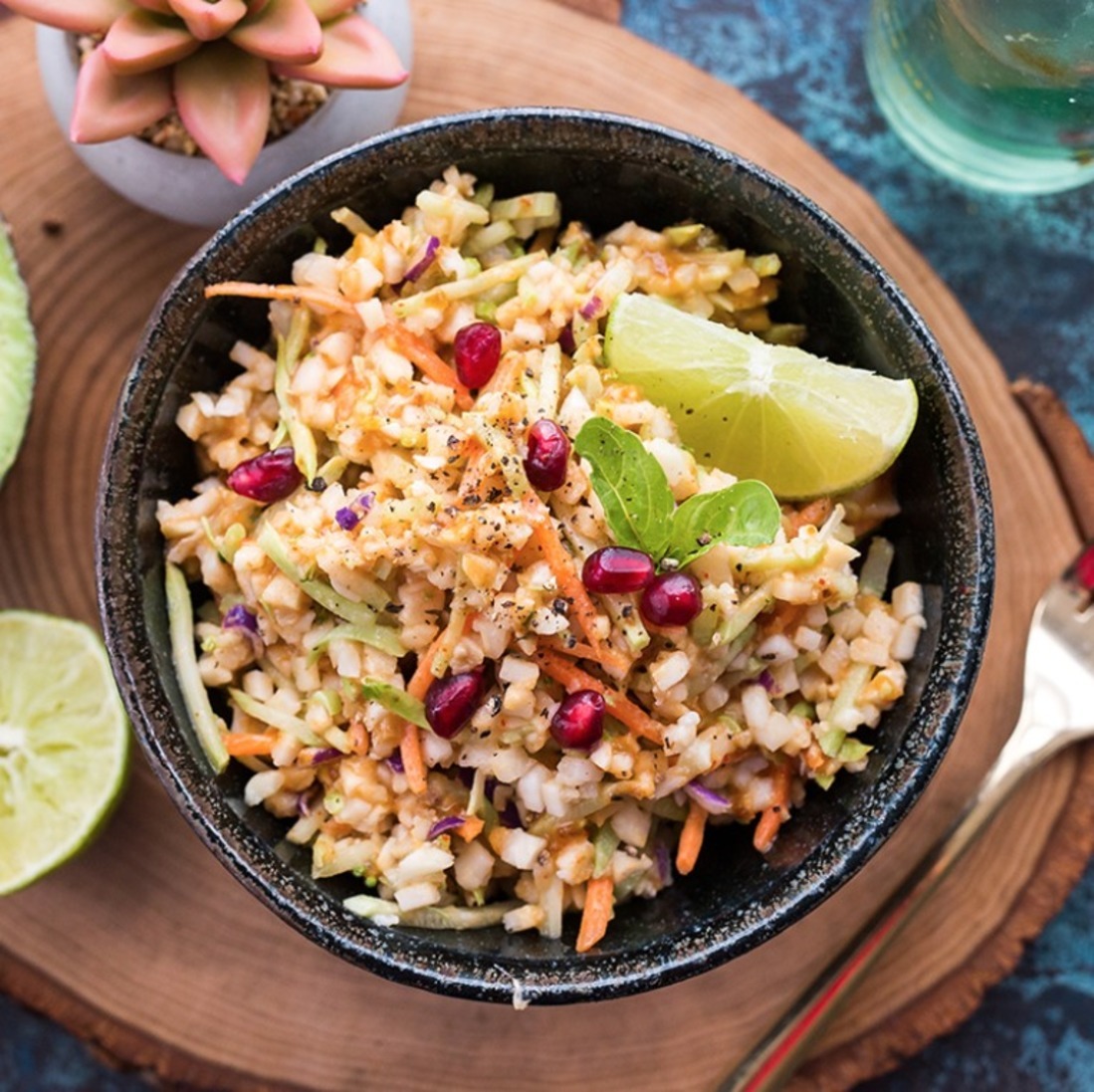 Raw veggie salad in a bowl with lime wedges and pomegranate seeds, on a blue textured surface.