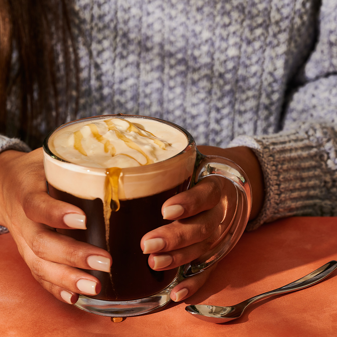 Hands holding clear mug of coffee with honey drizzle.