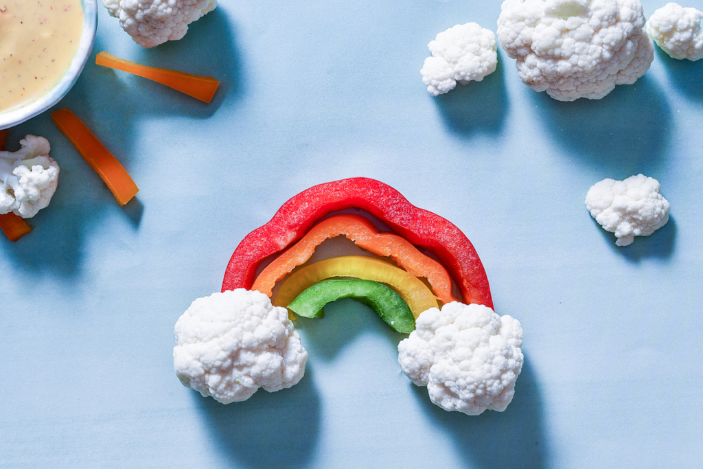 Rainbow-shaped bell pepper slices on a blue background with cauliflower clouds and carrot sunshine.