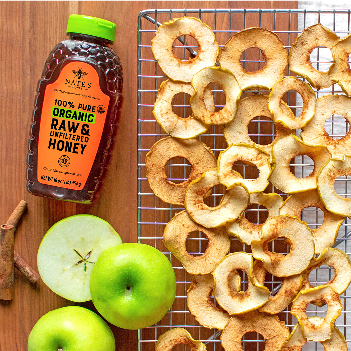 Nate's Organic Honey next to cooling rack with dried apple slices, fresh apple and cinnamon sticks on wood background