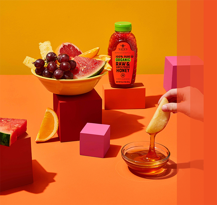 bowl of fruit next to bottle of Nate's Organic Honey on colored blocks on orange background.