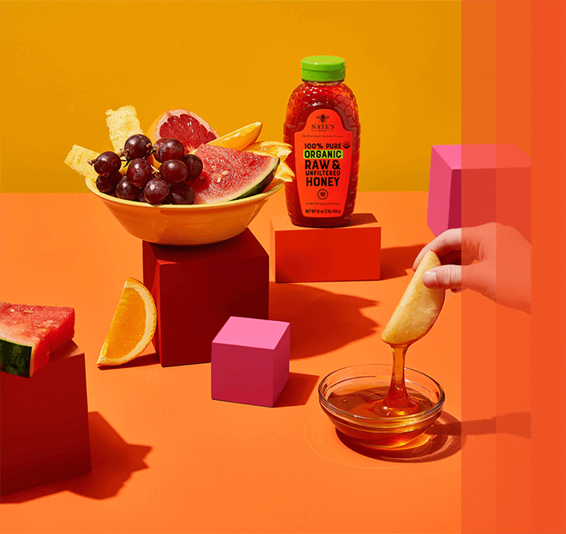 bowl of fruit next to bottle of Nate's Organic Honey on colored blocks on orange background.