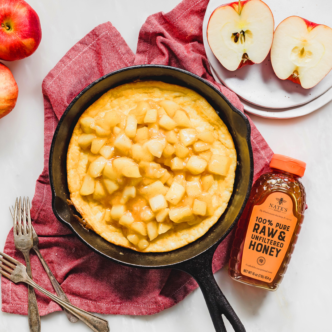 Skillet with a apple honey frittata, a bottle of Nate's Honey, sliced apples, burgundy cloth backdrop.