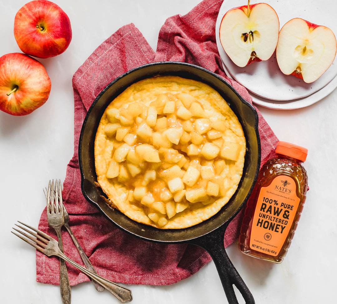 Skillet with a apple honey frittata, a bottle of Nate's Honey, sliced apples, burgundy cloth backdrop.