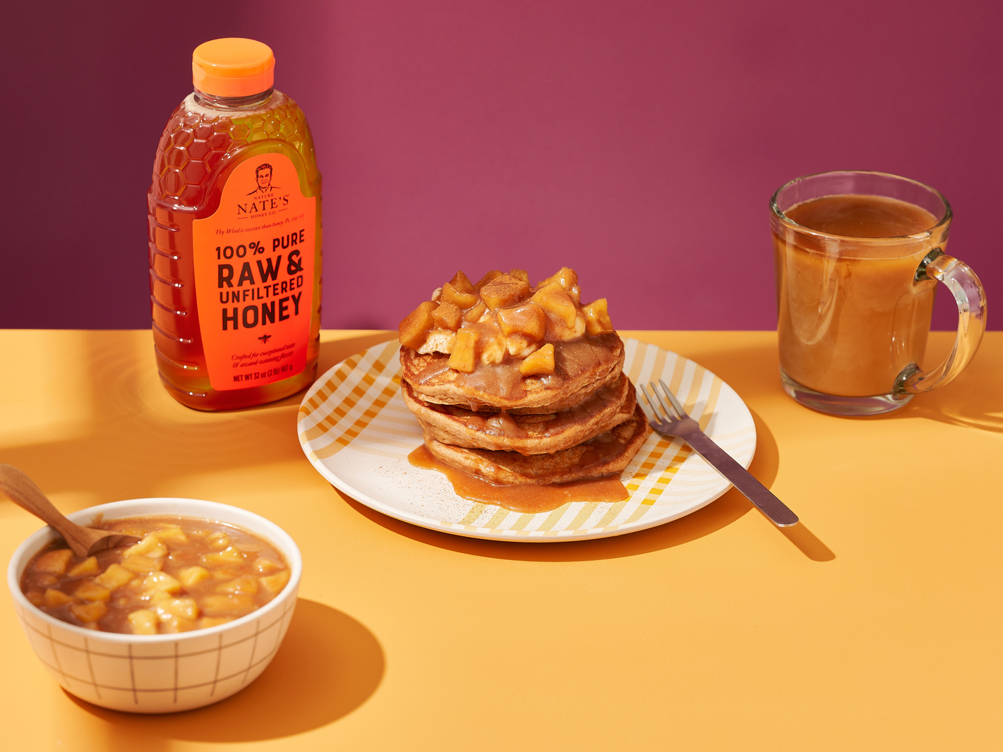 Nate’s honey bottle with a stack of apple pie pancakes and a mug of tea on a yellow table.