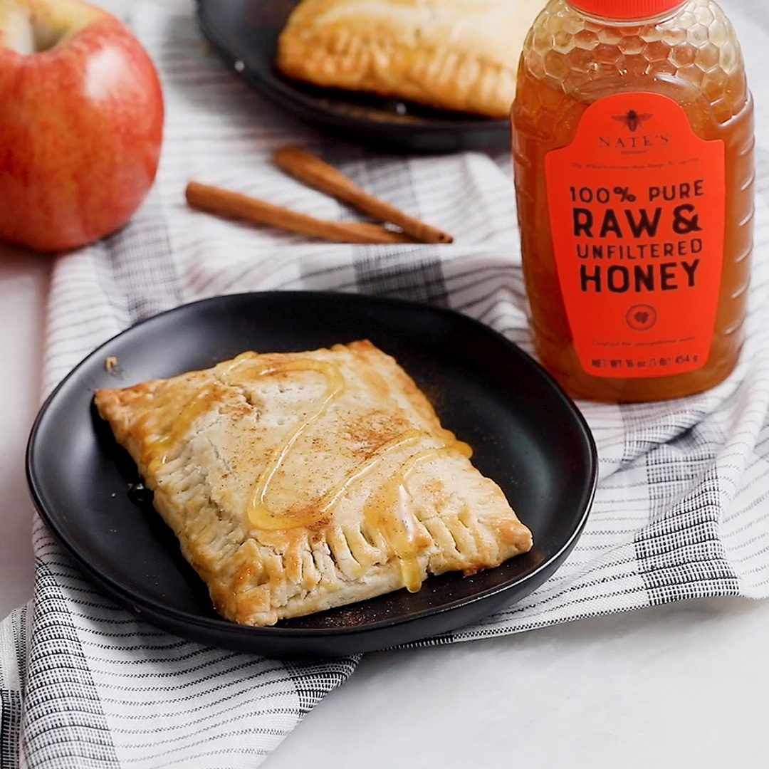 Apple turnover drizzled with honey on a plate with a bottle of Nate’s Honey and cinnamon sticks on a striped cloth.