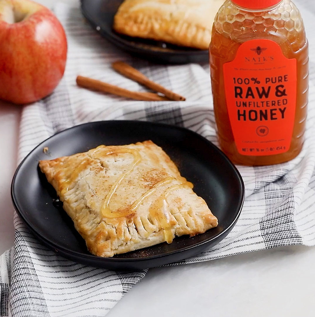 Apple turnover drizzled with honey on a plate with a bottle of Nate’s Honey and cinnamon sticks on a striped cloth.