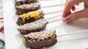 Chocolate-dipped apple slices with colorful sprinkles on a white plate, a person's hand seen picking one up.