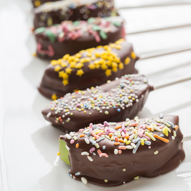 Chocolate-covered apples slices on sticks with colorful sprinkles on a white tray.