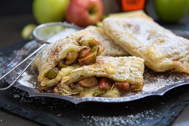 Apple and brie hand pies on a dark slate, fresh apples and Nate's honey bottle in background.