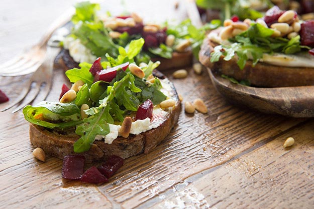 Toast topped with goat cheese, arugula, beets and pine nuts, drizzled with honey on a rustic wooden table.
