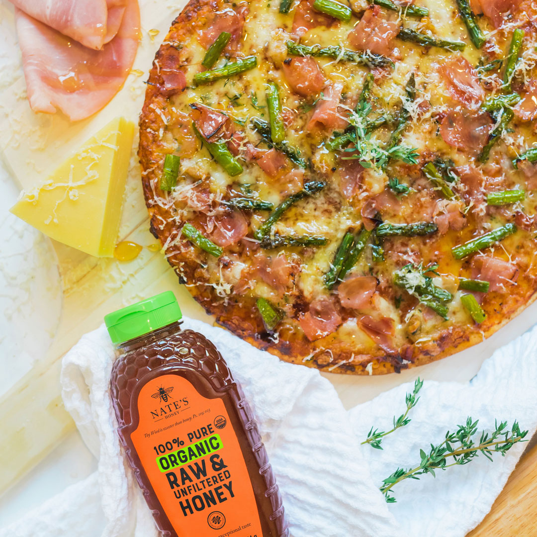 Asparagus and ham pizza with a bottle of organic Nate's Honey and cheese on a white crinkled cloth.