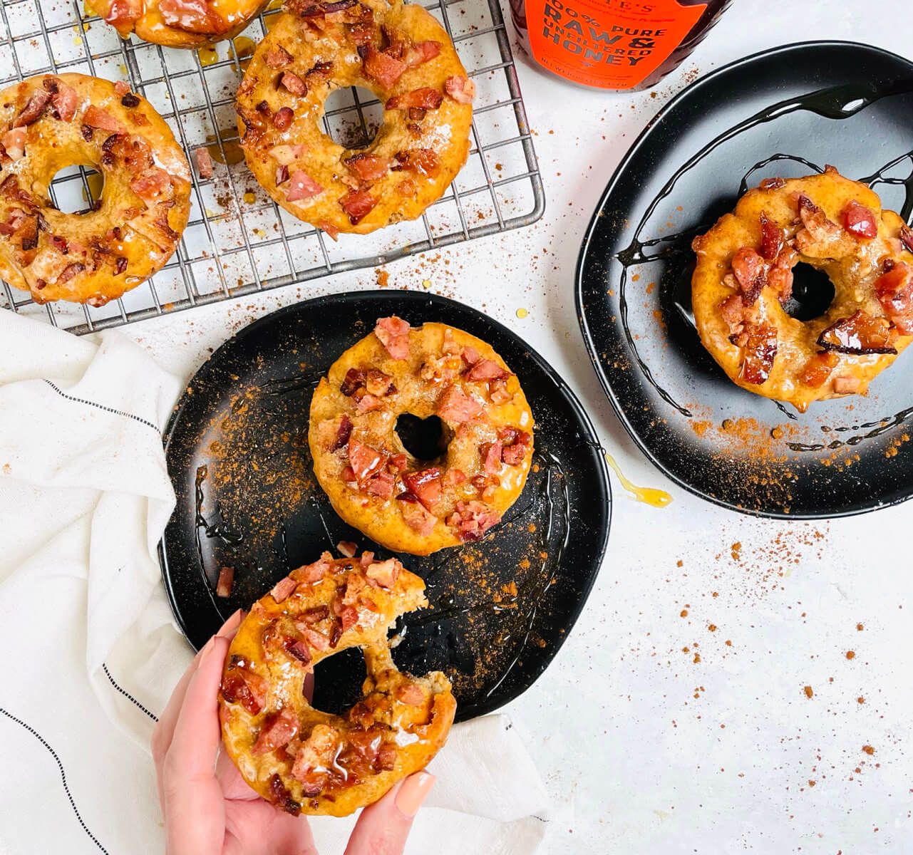 Donuts with bacon bits, drizzled with honey, on a wire rack and black plates, with a Nate's honey bottle on the side.