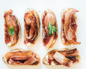 Assorted toasts topped with fruit spread and crispy bacon with fresh mint garnish displayed on a clean white background.