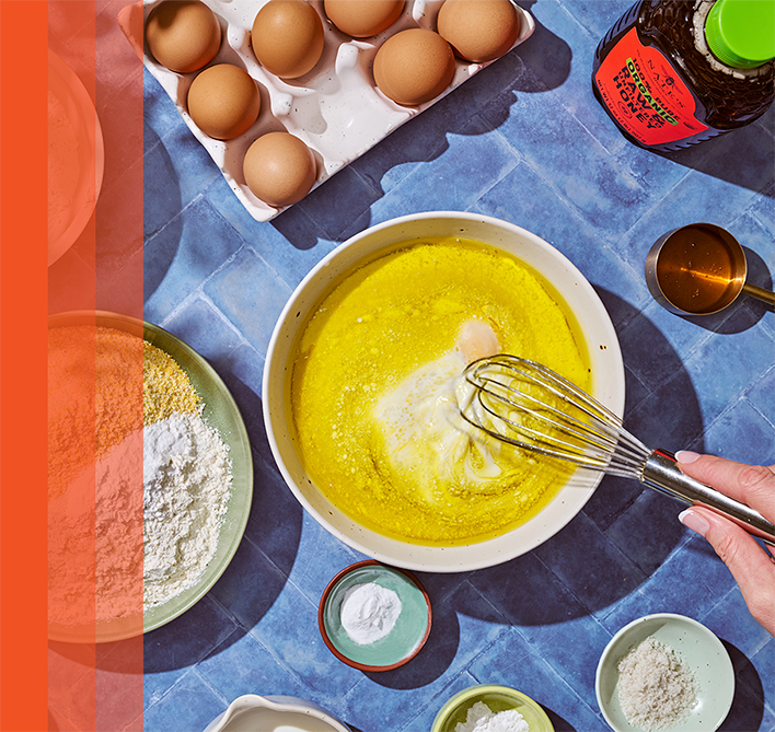 Bowl of yellow batter being whisked on a blue surface, surrounded by eggs, flour, and small bowls with a bottle of honey nearby.