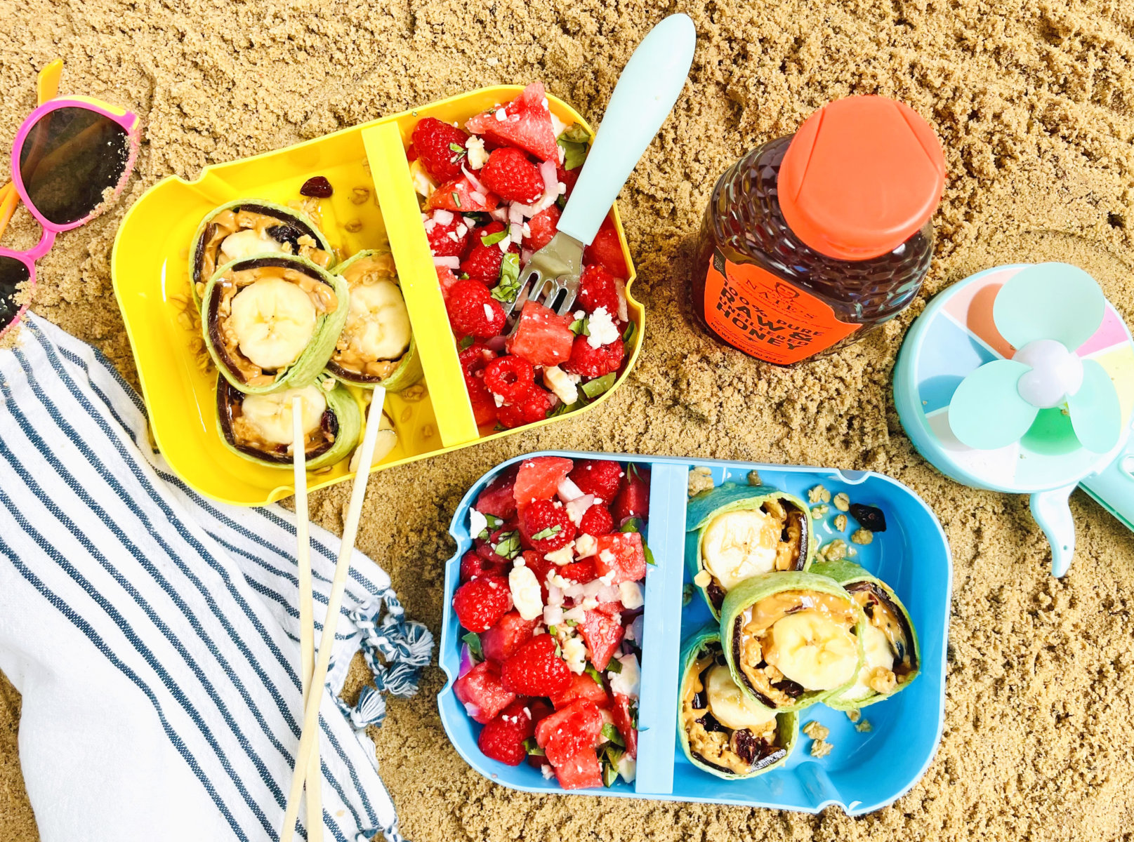 A beach picnic with fruit salads, banana roll ups, a bottle of Nate’s honey, and a small fan.