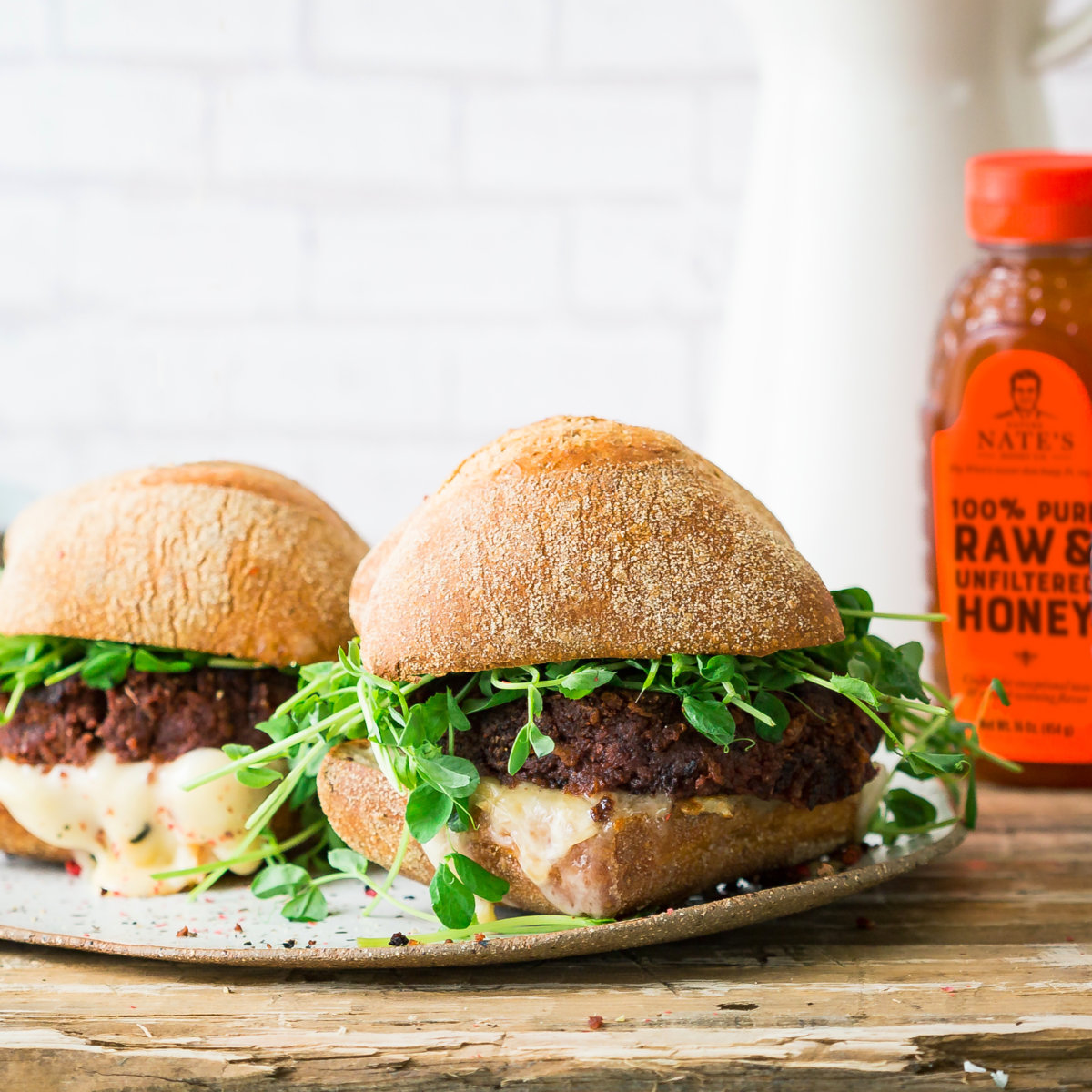 Two gourmet beet burger sandwiches with dark, crispy crusts, fresh green sprouts on top, and a bottle of Nate's honey.