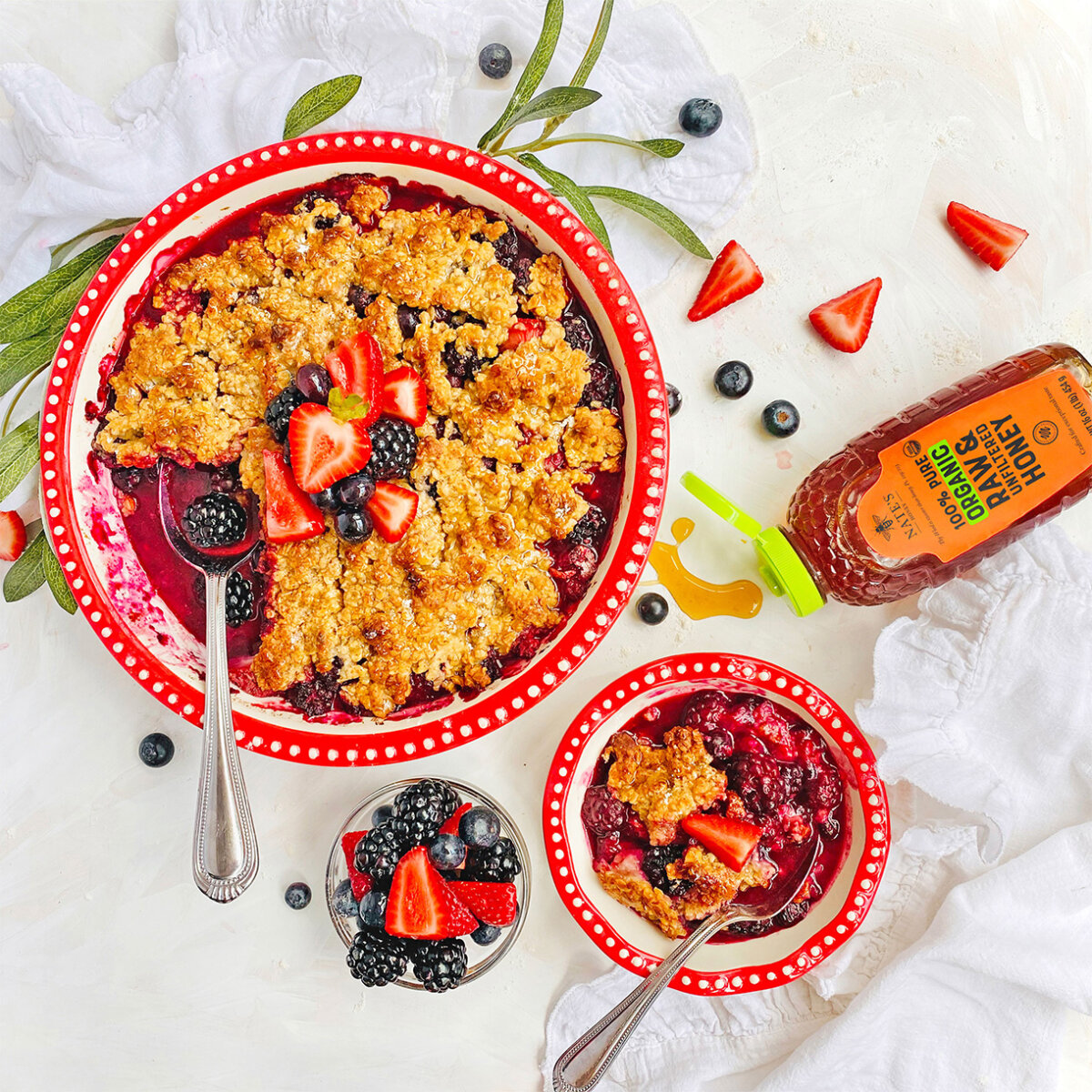 Red border platter with honey berry crumble next to ingredients, bowl of crumble and bottle of Nate's Organic Honey on white kitchen background.