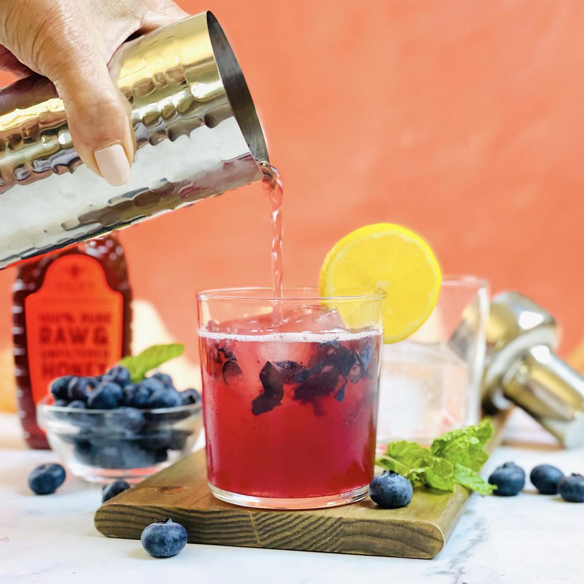 Hand pouring a berry cocktail with blueberries and a lemon slice garnish beside a bottle of Nate's Honey.