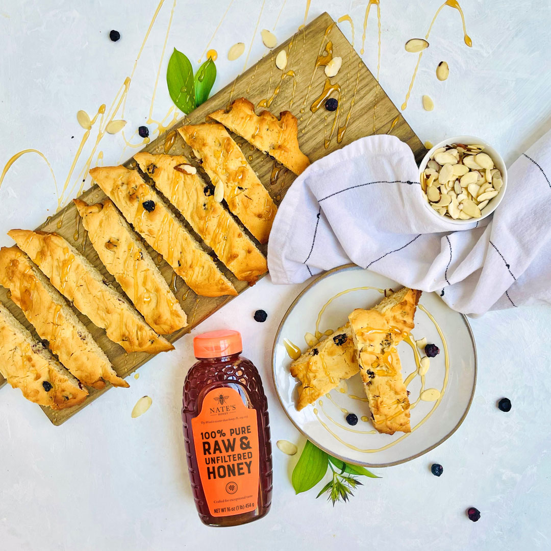 Golden biscotti and almonds on a board, drizzled with honey, with a bottle of Nate’s Honey and a blue background.