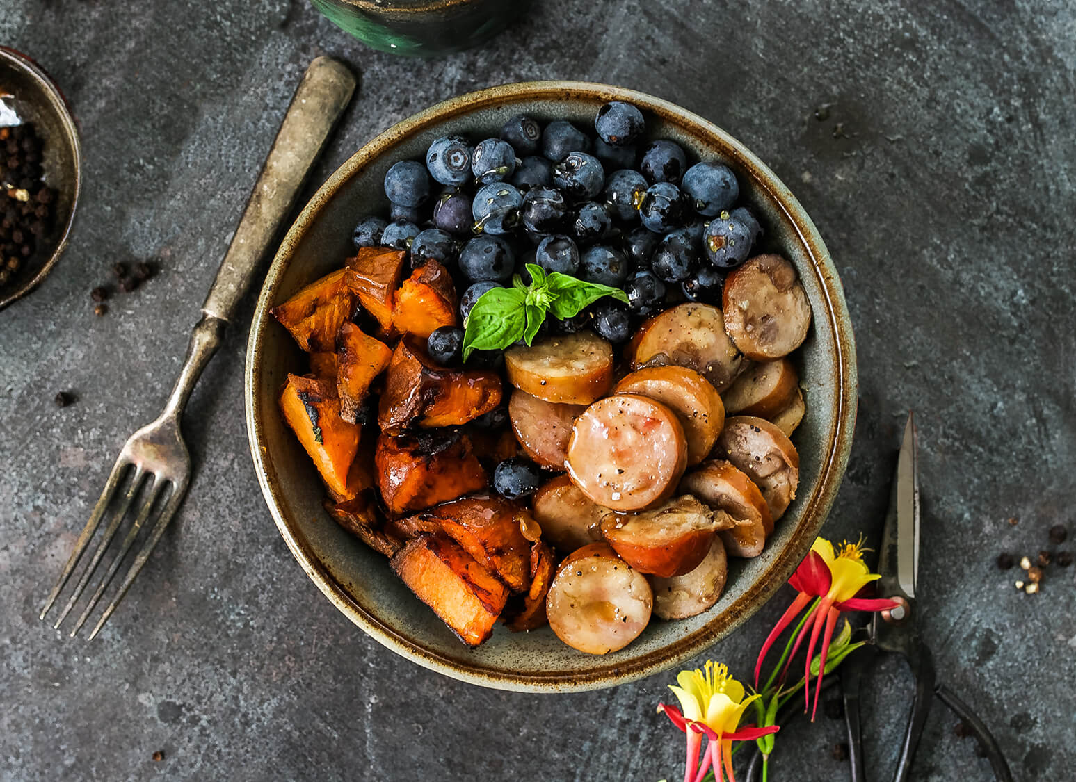A rustic dish with blueberries, sweet potatoes, sausage and basil on a dark surface, ready to eat.